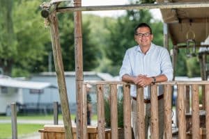 Mann lehnt sich an ein Holzgelände und lächelt in die Kamera. Im Hintergrund Bäume und ein Wohnwagen. 