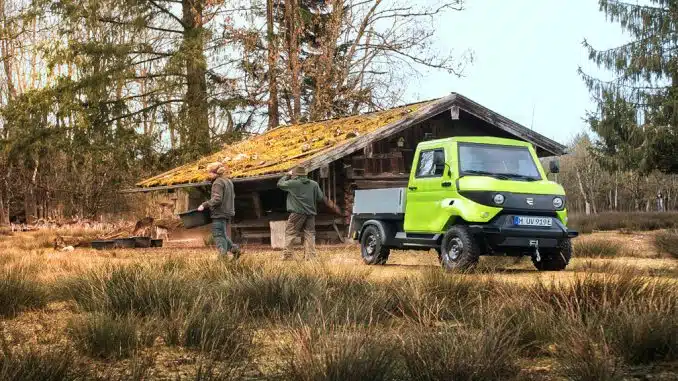 Grünes Elektro Auto mit Ladefläche in einem Wald vor einer Holzhütte.