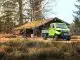 Grünes Elektro Auto mit Ladefläche in einem Wald vor einer Holzhütte.