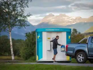 Mann der aus einem weiß-blauem Propangas-Automaten eine Gasflasche heraus in sein graues Auto hebt. Im Im Freien umgeben von Bäumen und Bergen im Hintergrund.