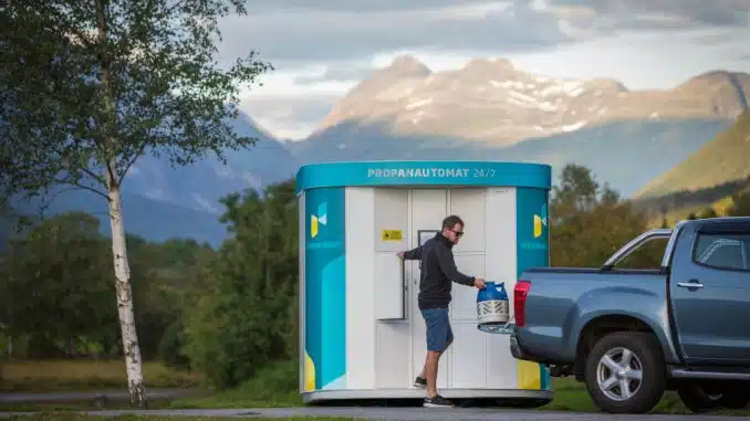 Mann der aus einem weiß-blauem Propangas-Automaten eine Gasflasche heraus in sein graues Auto hebt. Im Im Freien umgeben von Bäumen und Bergen im Hintergrund.