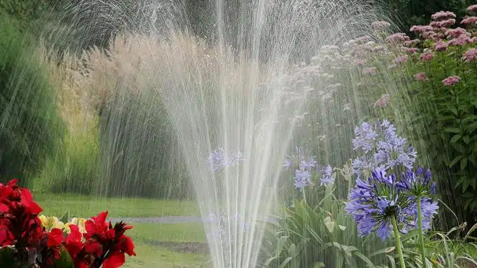 Garten grün mit roten und lilafarbenen Blumen. In der Mitte eine Wasserbewässerung.