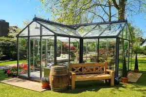 Gartenhaus aus Glas mit einer Bank und einem Bierfass davor in einem grünen, blühenden Garten mit Baum.