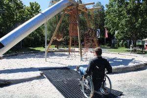 Mann im Rollstuhl auf einer Inklusionsmatte auf einem Spielplatz mit Kies und Klettergerüst.