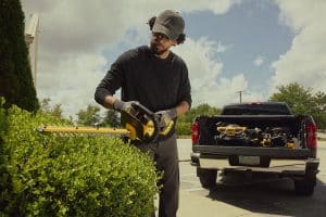 Mann mit DeWalt gelbe Heckenschere, der eine grüne Hecke scherrt. Im Hintergrund ist ein Auto zu sehen.