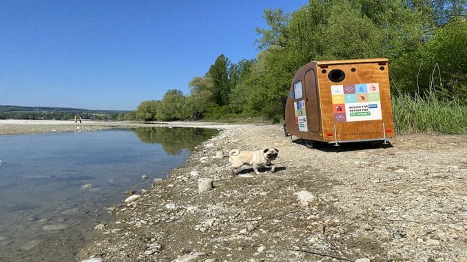 Fahrradanhänger für die EU Ecolabel Roadshow, aus Holz, an einem Ufer am See. Ein Hund im Vordergrund und Bäume im Hintergrund.