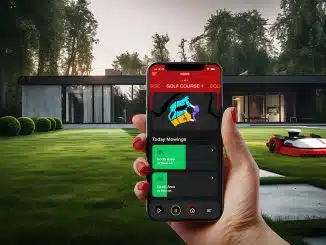 Smarter Mähroboter von Kress auf einem Rasen vor einem Haus. Im Vordergrund eine Hand mit Handy, die das Bedienmenü vom Mähroboter zeigt.