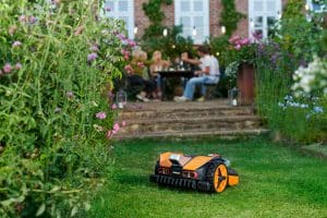 Smarter Mähroboter von Worx in einem Garten. Im Hintergrund eine Terrasse auf der Menschen feiern.